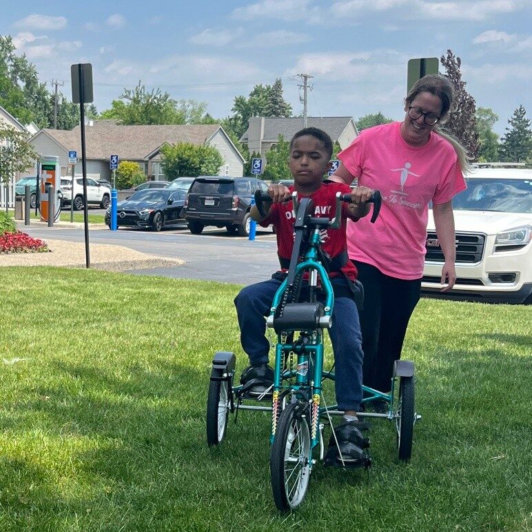 A young patient riding an adaptive tricycle outdoors with their therapist cheering them on.